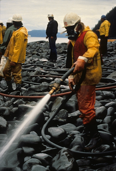 Crews wearing gas masks for protection wash down heavily oiled rocks on a shoreline