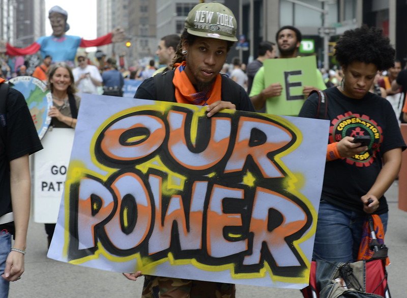 A young man of color holds a sign reading "Our Power" in big letters at a march through New York City. Next to him is a woman in a Cooperation Jackson t-shirt.